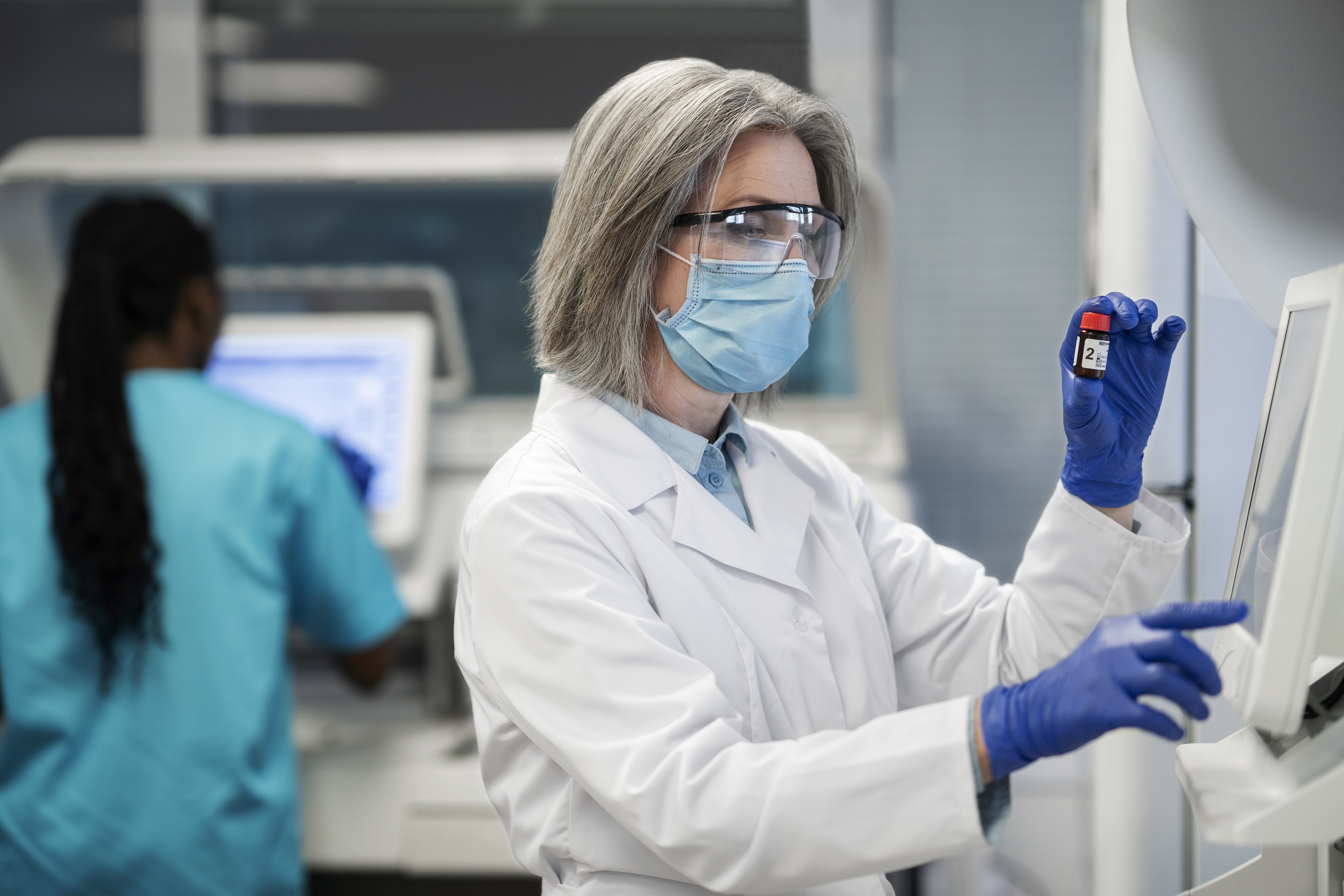 Médica de jaleco branco, máscara cirúrgica e luvas azuis analisa frasco de amostra em laboratório clínico